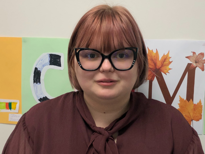 Alex, a young person looks at the camera. They have chestnut coloured bobbed hair with a fringe, and are wearing big glasses with black frames that have some sparkles on them and a brown blouse with a bow at the neck.