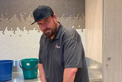 Chris, a young man with a beard looks to the camera as he stands in a wet area and holds cleaning equipment.