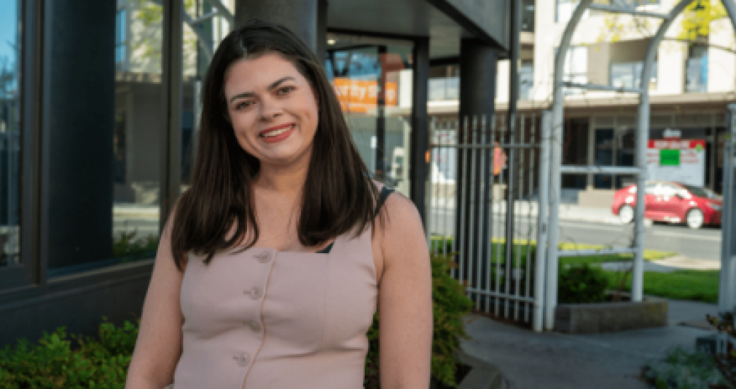 A smiling woman in a light peach sleeveless top, standing outdoors with buildings, greenery, and a red car in the blurred background.