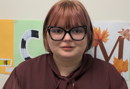Alex, a young person looks at the camera. They have chestnut coloured bobbed hair with a fringe, and are wearing big glasses with black frames that have some sparkles on them and a brown blouse with a bow at the neck.