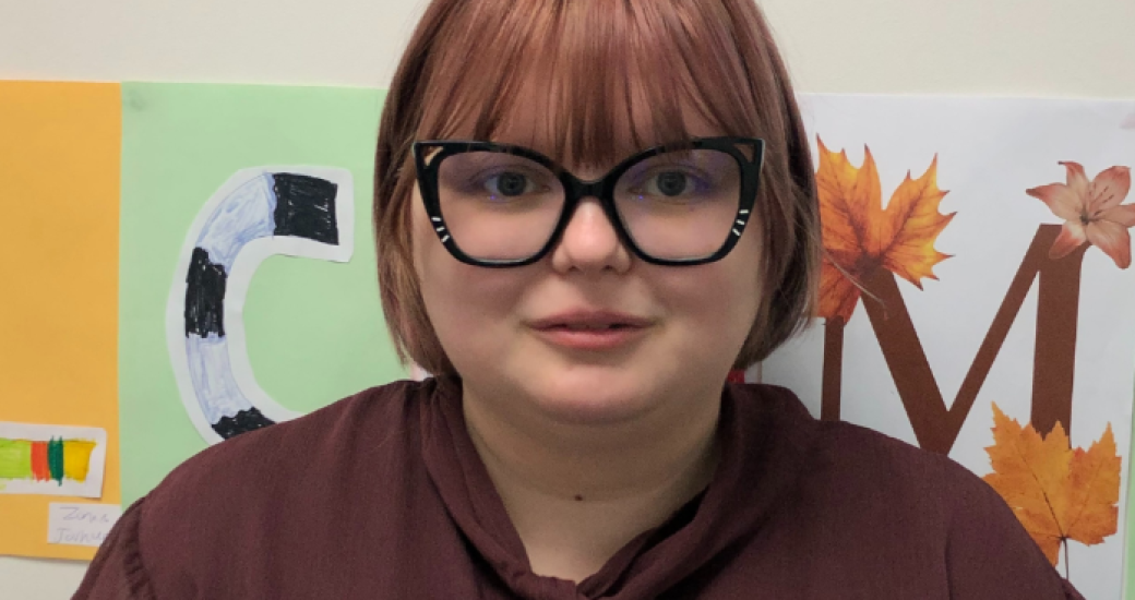 Alex, a young person looks at the camera. They have chestnut coloured bobbed hair with a fringe, and are wearing big glasses with black frames that have some sparkles on them and a brown blouse with a bow at the neck.