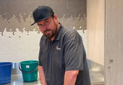 Chris, a young man with a beard looks to the camera as he stands in a wet area and holds cleaning equipment.