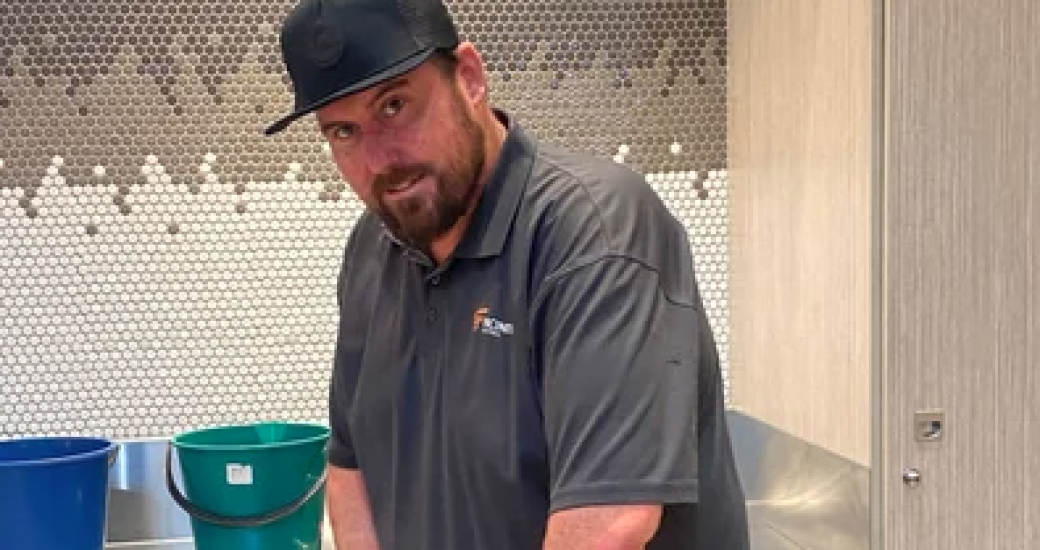 Chris, a young man with a beard looks to the camera as he stands in a wet area and holds cleaning equipment.
